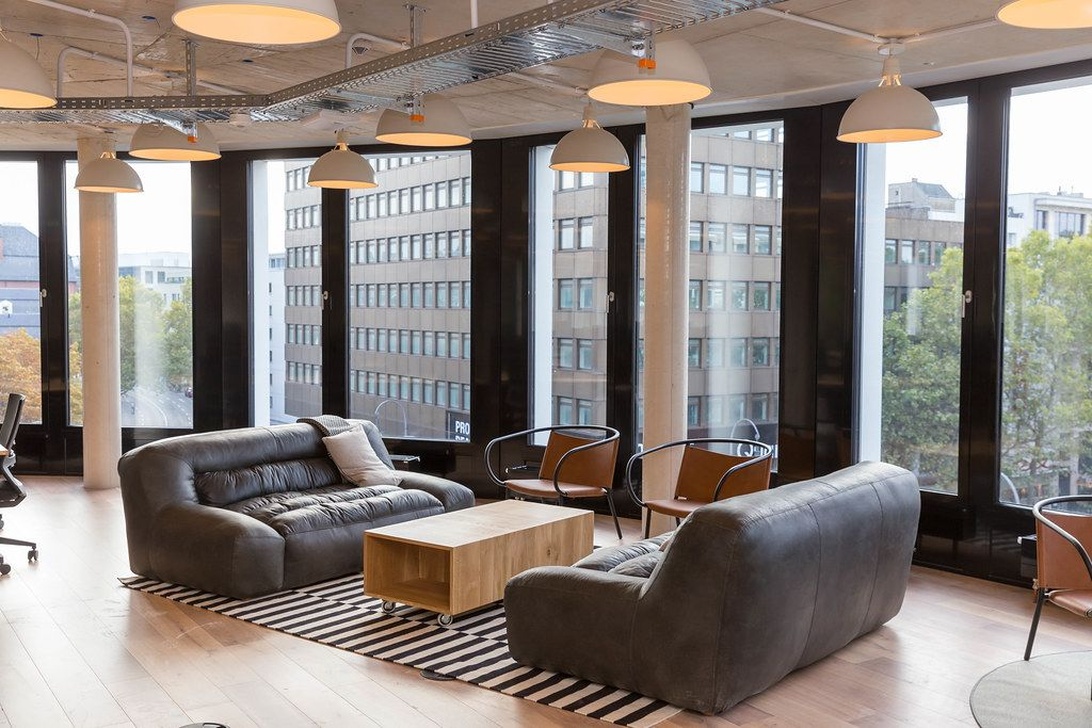 En modern, exklusiv coworking-lounge med fönster från golv till tak med utsikt över en stadssiluett. Utrymmet har bekväma grå soffor, soffbord i trä, designerbelysning och en ren estetik.