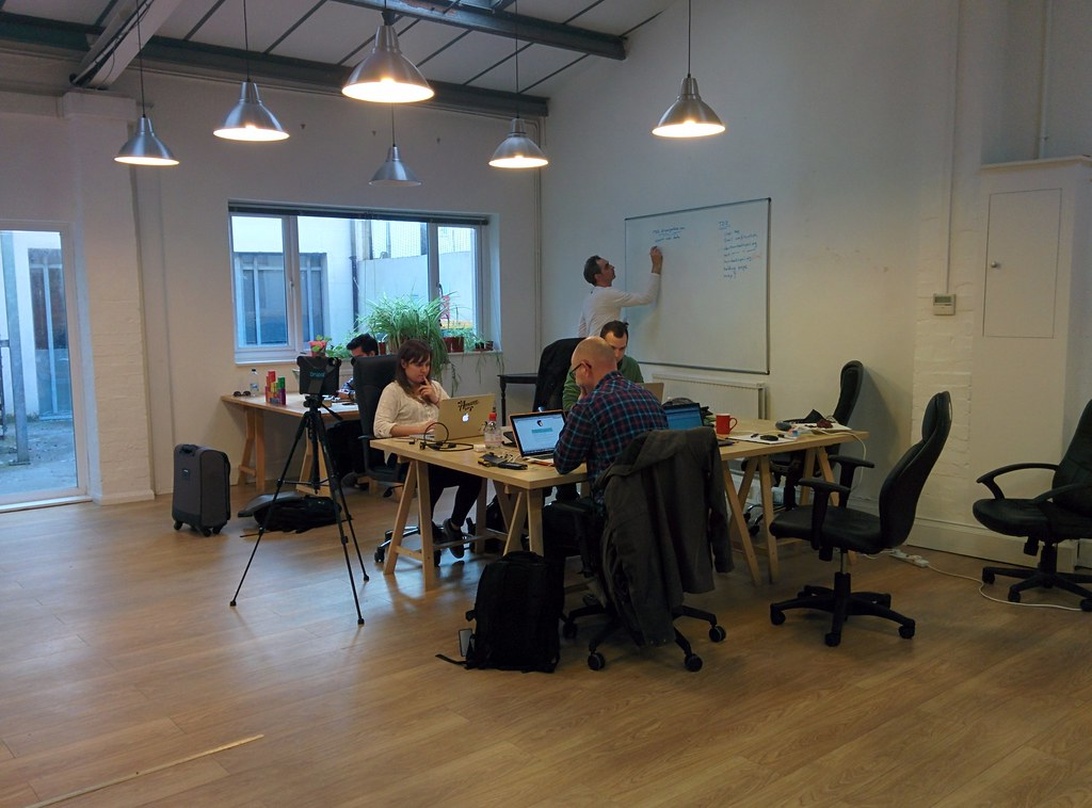 Ett coworking-utrymme med flera personer som arbetar vid träbord med bärbara datorer medan en person skriver på en whiteboardtavla. Utrymmet i industriell stil har taklampor och stora fönster.
